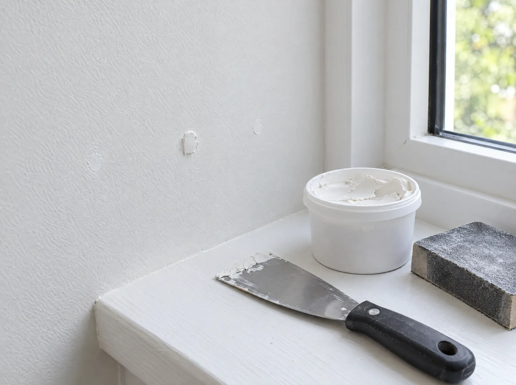 Frisch gespachtelte Dübellöcher an einer weissen Wand, daneben Spachtel und Schleifklotz auf dem Fenstersims